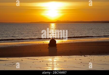 Portobello, Edinburgh, Schottland, Großbritannien. 5.. April 2020. Sonnenaufgang am Strand von Portobello, mit einer Frau, die am Sandstrand in Kontemplation sitzt, und verschiedenen anderen Menschen, die Schwimmen, Wandern, Laufen fotografiert haben. Stockfoto