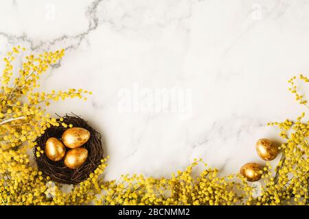 Goldene Wachteleier in kleinem Gelege mit gelben bunten Mimosenblüten auf Marmortisch. Frohes Osterkonzept. Hintergrund der Draufsicht mit Kopierbereich Stockfoto