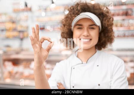 Nahaufnahme der fröhlichen Mädchen in der Nähe von Ladentisch posiert, zeigt ok Geste, frisches Fleisch Produkte im Kühlschrank. Porträt einer jungen lächelnden Frau mit Cur Stockfoto