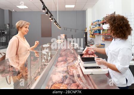 Seitenansicht der jungen lächelnden Frau mit lockigen Haaren demonstriert positive Dame rohes Schweinefleisch. Fröhliches Mädchen hinter Ladentisch mit großen Sortiment showi Stockfoto