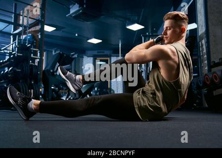 Muskulöser Kaukasusmann trainiert abs auf dem Boden im Sportverein. Seitenansicht des gutaussehenden jungen Sportlers Gebäude Kernmuskeln, tun knirscht in leerem Fitnessstudio. Stockfoto
