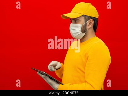 Lieferung Mann in gelben Hut, leeres T-Shirt in Form eines sterilen Gesichtsmaske Handschuhe, hält eine Tablette, isoliert auf rotem Hintergrund. Mitarbeiter arbeitet Stockfoto