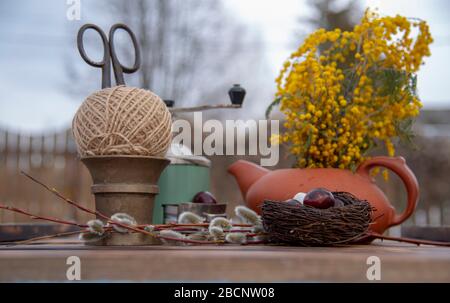 Auf der Holztischplatte befindet sich ein Gelege mit bemalten Wachteleiern, Weidenästen und verschiedenen Haushaltsgeräten. Stockfoto