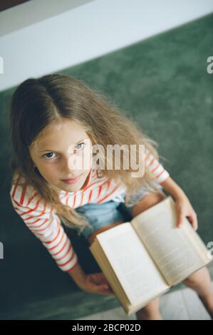 Cleveres kleines Mädchen mit langen Haaren, das auf dem grünen Sofa sitzt und Buch auf dem Knie hat Stockfoto