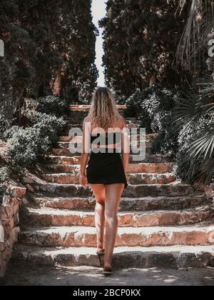 Blonde junge Frau, die an einem sonnigen Sommertag mit kleinen Kleidern eine Treppe mitten in der Natur klettert Stockfoto