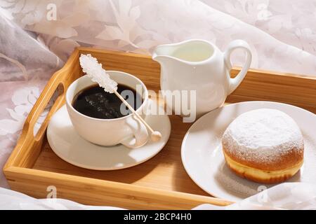 Frühstück im Bett mit frisch gebrühtem Kaffee, Kristallzucker, Milch und Donut in Puderzucker, auf einem Holztablett in weißen Gerichten. Stockfoto