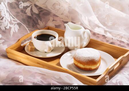 Frühstück im Bett mit frisch gebrühtem und aromatischem Kaffee, Rohrzucker, Krug mit kalter Milch und Donut in Puderzucker auf einem Holztablett in weißen Gerichten Stockfoto