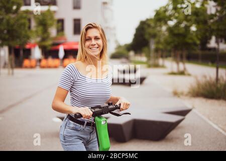 Hübsche junge Frau ist in der Stadt mit einem Elektroroller unterwegs Stockfoto