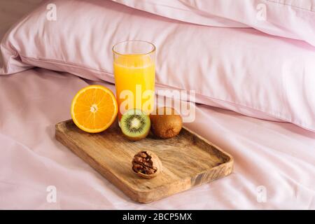 Morgens gesundes Frühstück im Bett mit frisch gepresstem Orangensaft, halber Orange, Kiwi und Walnuss auf einem Holztablett vor dem Hintergrund der rosa Kissen Stockfoto