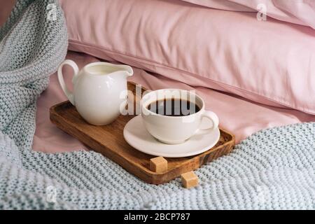 Frühstück im Bett mit frisch gebrühtem und köstlichem Kaffee, einer Kanne mit kalter Milch und Rohrzucker auf einem Holztablett vor dem Hintergrund der rosa Laken, pillo Stockfoto