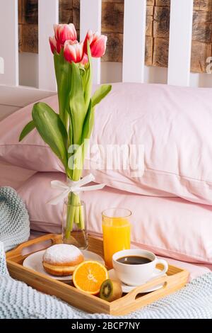 Frühstück im Bett mit einer Tasse frisch gebrühten Kaffee, Orangensaft, Orange, süßer Kiwi, Donut und einem Bouquet von frisch geschnittenen Tulpen in Vase in einem Holz Stockfoto