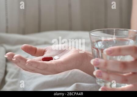 Nahaufnahme der Hände junger Frau. Mädchen im Bett, das Tabletten oder Pillen und Glas Wasser hält. Painkiller mit Platz für Text. Verschwommener Hintergrund. Stockfoto