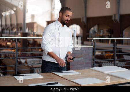 Hübscher junger Koch Afrikanische Ethnie legt Messer auf dem Tisch vor einem kulinarischen Master Class. Afro Mann in Chef einheitliche Fertig, für das Kochen von Cla Stockfoto