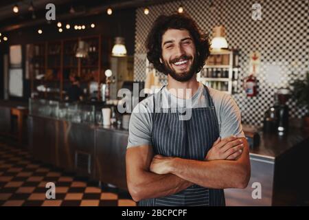 Portrait eines lächelnden Kellners, der im Café mit gefalteten Händen steht Stockfoto