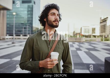 Junger Geschäftsmann, der im Freien eine Tasse Kaffee zum Mitnehmen hält, während er wegschaut und denkt Stockfoto