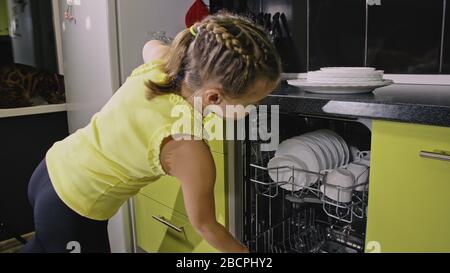 Schlaue Mädchen lernen, Geschirrspüler zu benutzen. Stilvolle, moderne Einbauküche in grünem Schwarz. Kind legt sauberes Geschirr. Stockfoto