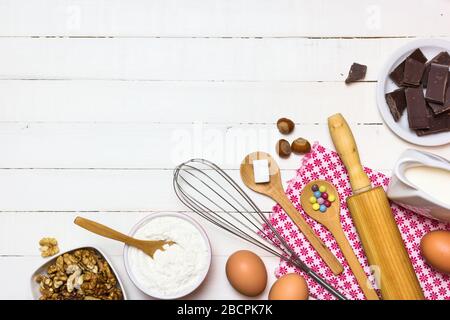 Zutaten und Utensilien zum Backen von Kuchen. Eier, Mehl, Milch, Zucker, Nüsse und Schokolade auf weißem Holztisch. Backen- oder Kochkonzept. Stockfoto