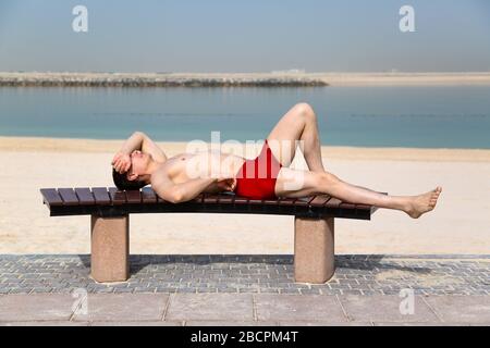 Ein junger Kerl in roten Badestämmen sonnt sich auf einer Bank am Strand. Stockfoto