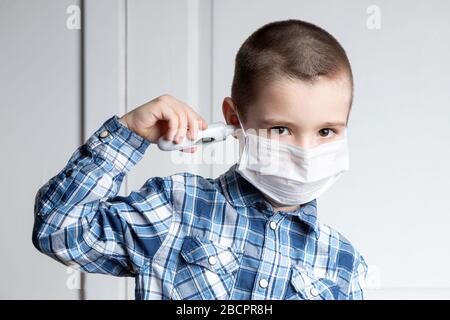 Der junge Junge mit schmerzenden Augen in einer medizinischen Maske misst die Temperatur eines Thermometers im Ohr. Medizinischer Schutz vor durch die Luft übertragenen Krankheiten, Koronav Stockfoto