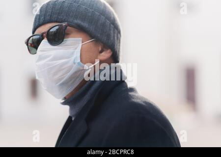 Bild von erwachsenen Mann trägt Schatten und schützende medizinische Maske, Spaziergänge im Freien während Coronavirus-Epidemie, sieht beiseite, denkt, wie Virus zu überwinden, blu Stockfoto