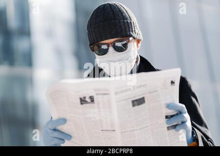 Foto des europäischen Mannes trägt schützende medizinische Maske und Handschuhe, Spaziergänge durch die Stadt im Freien, liest Artikel über Virenausbruch in verschiedenen Ländern Stockfoto