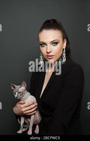 Modeporträt der eleganten Luxusfrau in Rautenohrringen mit sphynx-katze auf Schwarz Stockfoto