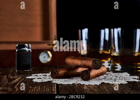 Stillleben mit drei kubanischen Zigarren, zwei Gläsern Whisky oder Rum, leichtere und Holzkiste mit Hygrometer. Alte Holztischplatte und schwarzer Hintergrund. Stockfoto