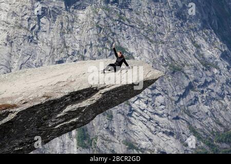 Furchtlose Frau, die auf der Trolltunga-Felsformation sitzt und die Hand winkt. Die Felswand befindet sich in Odda, Bezirk Hordaland, Norwegen Stockfoto