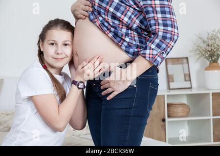 Überraschte Teenager-Tochter, die Baby im Bauch ihrer schwangeren Mutter hört und mit Händen berührt, glückliches Mädchen, das Kamera betrachtet Stockfoto