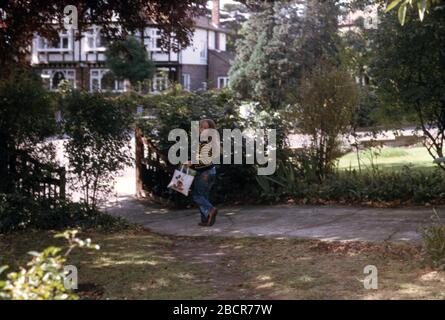 Ein junges Mädchen, das einen gelb-blau gestreiften bretonischen Pullover und eine blau gestreifte Jeans trägt und eine Micky-Maus-Tasche mit Geschenken im Vorgarten ihres Familienhauses, Großbritannien 1974, trägt Stockfoto