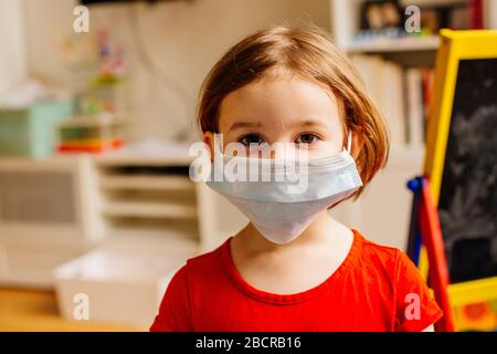 Portrait eines kleinen Kindes mit Virusschutz chirurgische Gesichtsmaske, die in ihrem Raum für soziale Distanzierung aufgrund von Coronavirus zuhause bleibt Stockfoto