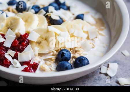 Bircher Müsli mit Beeren, Bananen, Marmelade und Kokosnuss in einer weißen Schüssel, grauem Hintergrund. Stockfoto