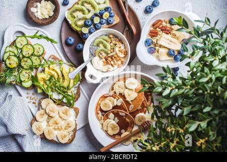 Gesunder, abwechslungsreicher vegetarischer Frühstückstisch. Haferbrei mit Obst, chia Pudding, Pfannkuchen mit Bananen und Honig und Toast mit Obst, Gemüse und cr Stockfoto