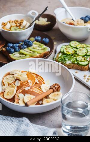 Gesunder, abwechslungsreicher vegetarischer Frühstückstisch. Haferbrei mit Obst, chia Pudding, Pfannkuchen mit Bananen und Honig und Toast mit Obst, Gemüse und cr Stockfoto