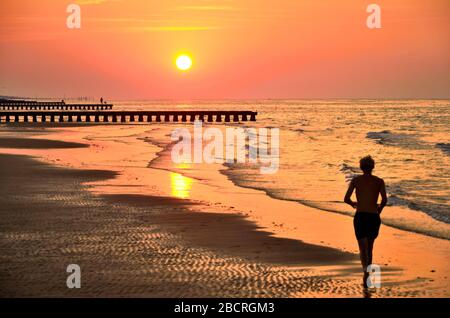 Silhouette des jungen Mannes, der bei Sonnenaufgang auf Strandsand läuft. Originaltapete aus dem aktiven Sommerurlaub. Stockfoto