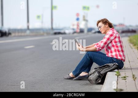 Frau im mittleren Alter beginnt auf den Straßen der Stadt mit dem Bloggen, indem sie Smartphone verwendet und am Straßenrand sitzt Stockfoto