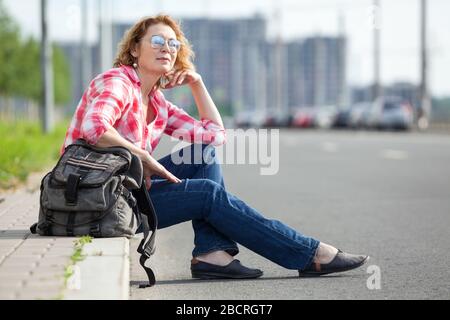 Kaukasische Frau fährt auf leerer Asphaltstraße, allein Reisende sitzen am Straßenrand und warten auf das Auto Stockfoto