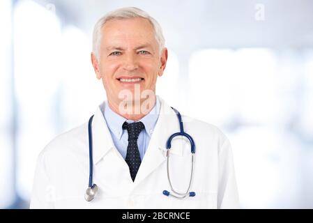 Portraitaufnahme eines älteren männlichen Arztes, der die Kamera betrachtet und lächelt, während er im Krankenhaus steht. Stockfoto