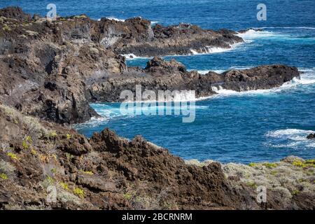 Vulkanische Felsformation, Klippen aus schwarzer Lava am felsigen Ufer mit zerdrückenden weißen Wellen über dem Atlantik. Blauer Himmel Hintergrund. La Palma, Can Stockfoto