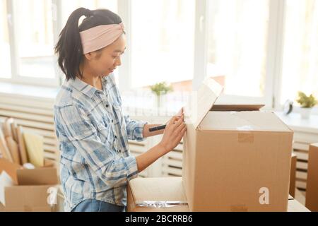 Seitenansicht Porträt der jungen Asiatin, die Kartons bei Sonneneinstrahlung unterschreibt, während sie in ein neues Haus oder eine neue Wohnung ausziehen, Kopierraum Stockfoto