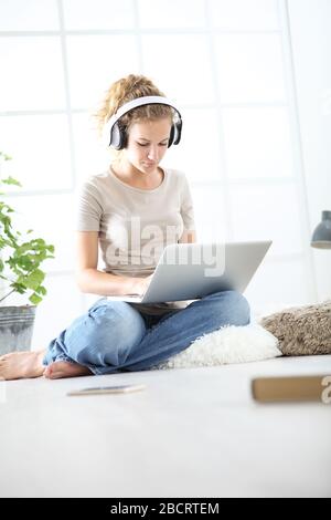 Junge Frau, die im Wohnzimmer sitzt und mit Kopfhörern und Computer studiert, bleibt beim Konzept zu Hause Stockfoto