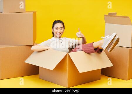 Lange Portrait von glücklicher Asiatin, die in der Box sitzt und Daumen nach oben zeigt, Studio auf hellgelbem Hintergrund, Kopierraum gedreht Stockfoto
