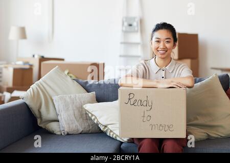 Porträt der jungen asiatischen Frau, die Kartonschachtel mit fahrbereiter Aufschrift hält und lächelnd mit Blick auf die Kamera blickt, Kopieraum Stockfoto