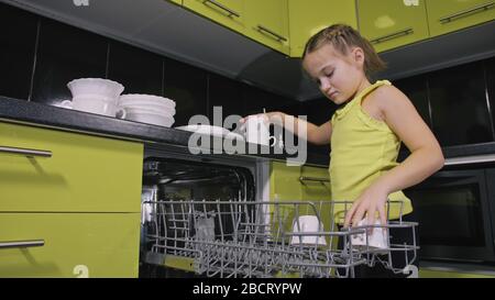 Schlaue Mädchen lernen, Geschirrspüler zu benutzen. Stilvolle, moderne Einbauküche in grünem Schwarz. Kind legt sauberes Geschirr. Stockfoto