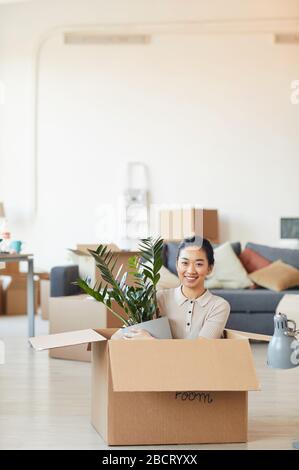 Vertikales Porträt der jungen asiatischen Frau, die in der Box sitzt und die Anlage hält, während sie in ein neues Haus oder eine neue Wohnung einzieht, kopiere Raum Stockfoto