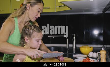 Mutter unterrichtet Tochter Smart Mädchen lernen zu kochen. Herrscherkinder zum Kochen eines neapolitanischen Eiergebratenen Omeletts aus salameimer Affumikatowurst. Stockfoto