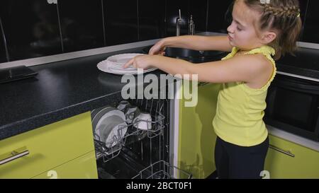 Schlaue Mädchen lernen, Geschirrspüler zu benutzen. Stilvolle, moderne Einbauküche in grünem Schwarz. Kind legt sauberes Geschirr. Stockfoto