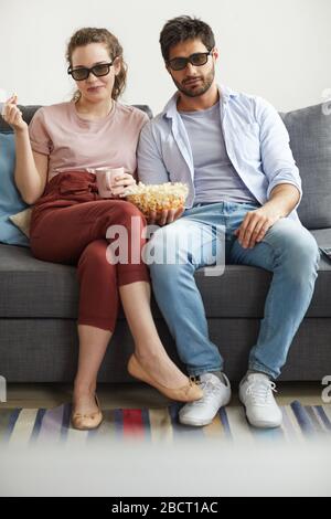 Vertikales, durchgehender Porträt eines modernen Paares, das Fernsehen zusammen sieht, während es auf dem Sofa sitzt und zu Hause Sonnenbrille trägt, und dabei die Faulheit genießt Stockfoto