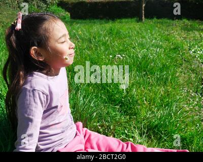 Entspannt, kleines Mädchen, das im Gras im Sonnenschein sitzt und eine Blume im Mund hält Stockfoto