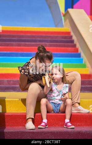 Junge Mutter mit Brille und Tochter sitzt auf einer farbigen Treppe. Kinderschutzkonzept, Muttertag Stockfoto